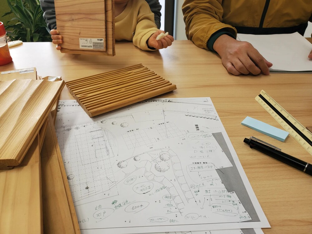 住宅の設計打ち合わせ風景。木材のサンプルや設計図面がテーブルに広げられ、大人と子どもが一緒に家づくりの素材選びをしている様子。