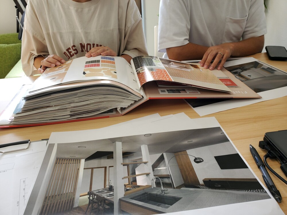 新築住宅の設計打ち合わせ風景。カタログやパースを囲み、ご夫婦が楽しげに素材や空間の検討をしている様子。建築家と住まい手が共に未来の暮らしを描いている場面。