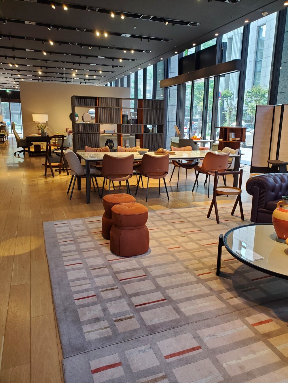 大きなガラス窓から自然光が差し込むショールームのインテリア展示。木目の床と幾何学模様のラグの上に、落ち着いた色合いのダイニングセットや革張りチェアが並び、和モダンとジャパンディを融合させたホテルライクな空間演出