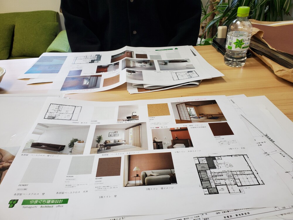 やまぐち建築設計室の設計打ち合わせ風景。新築住宅の間取り図とインテリアコーディネート資料を机に広げ、壁・床・天井の色や素材、照明との相性を比較検討している様子。色彩心理を踏まえ、暮らしの心地よさを整えるための設計プロセス。