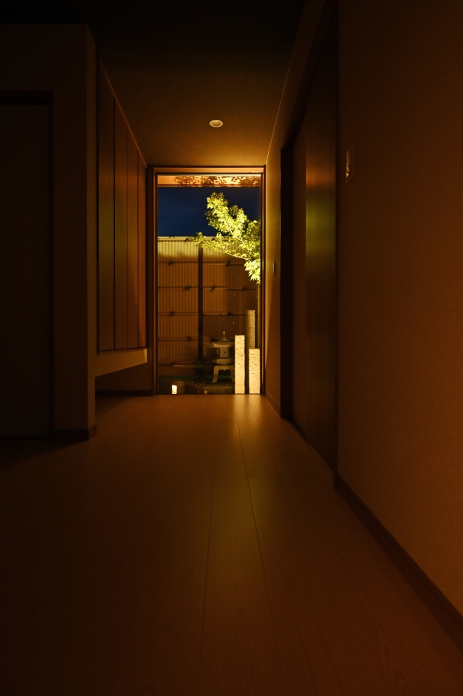 夜の玄関ホールから坪庭へと視線が抜ける、和モダン住宅の空間設計。廊下の突き当たりに設けた庭を大開口で切り取ることで、室内に奥行きと静けさを演出。柔らかな間接照明と植栽のライトアップが、旅館のような癒しとホテルライクな上質感を生み出している。視線の抜け、陰影、余白を丁寧に整えた設計により、帰宅した瞬間から心が整う住まいを実現。黒を基調とした外観、坪庭、和風建築、数寄屋、デザイナーズ住宅に憧れる方へ向けた、奈良県の建築家による注文住宅実例。和モダンの家、上質な空間、庭とつながる暮らし、静かな住環境を求める方におすすめの住宅デザイン。