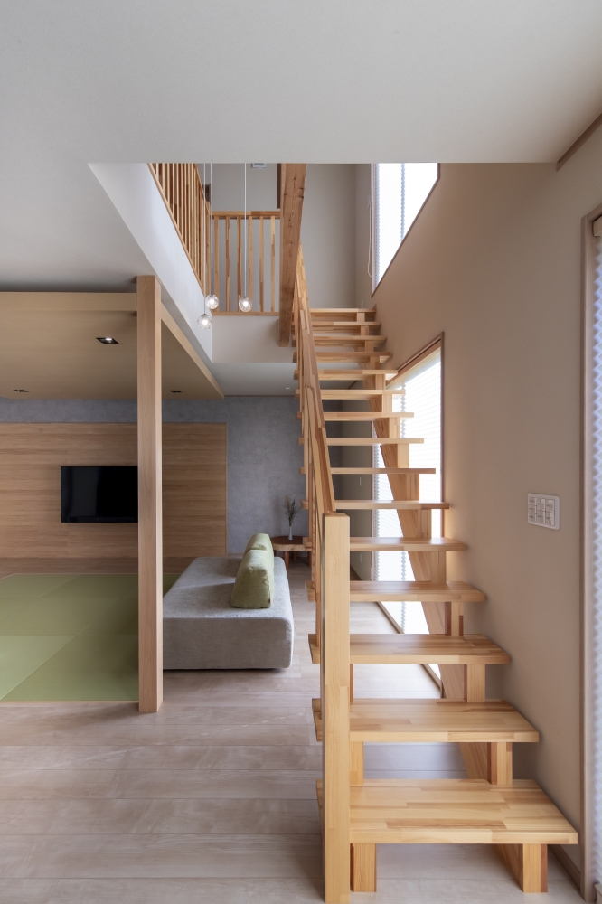 吹抜けと一体化した、和モダン住宅のリビングアクセス階段空間。無垢材を活かした軽やかなオープン階段と木格子の手すりが、空間に開放感と温もりを与え、ホテルライクで上質な暮らしを演出している。大きな窓から差し込む自然光と吹抜けの縦方向の広がりが、LDK全体に心地よい抜け感を生み出し、家族の気配を自然につなぐ設計。階段を単なる移動空間ではなく、腰掛けたり、会話が生まれたりする“居場所”として計画することで、日常の過ごし方に豊かさを加えている。和モダンの家・リビング階段・吹抜けリビング・高級注文住宅・デザイナーズ住宅・木の温もりを感じる家を求める方に向けた建築実例。