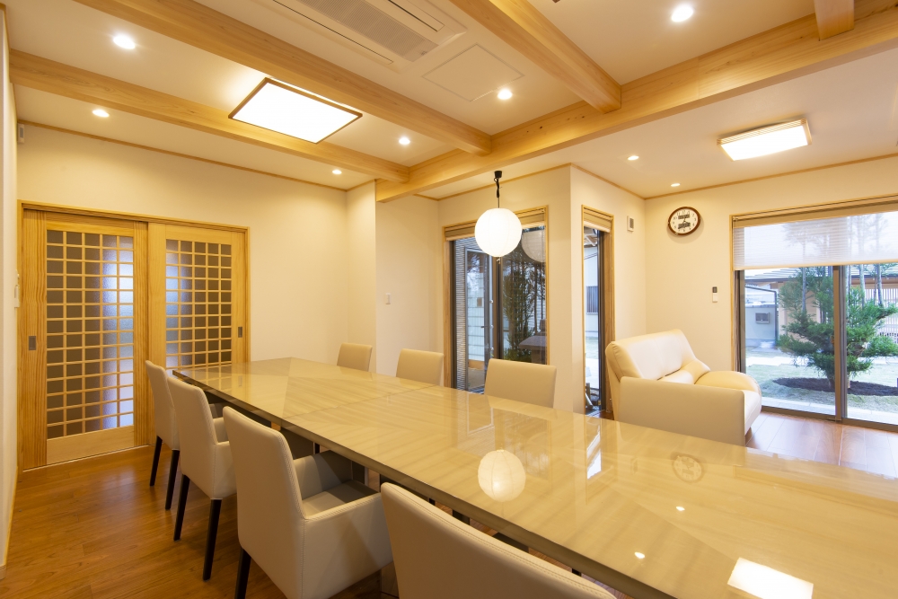 奈良県のやまぐち建築設計室が設計した和モダン住宅のダイニング・リビング一体空間。無垢材の梁を現した天井と上質な長尺ダイニングテーブルを中心に、障子格子の引き戸と大開口サッシにより庭と緩やかにつながる設計。自然光と間接照明が調和し、家族の団らんと来客対応を両立するホテルライクな高級注文住宅のLDK実例。