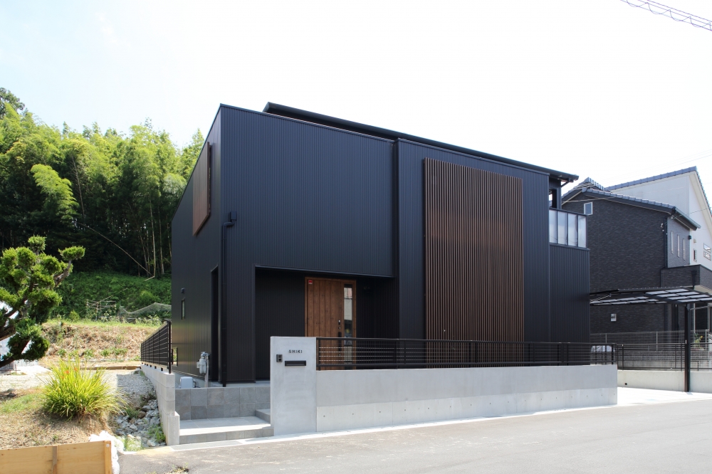 奈良県の住宅地に建つ和モダン住宅の外観。黒のガルバリウム鋼板と縦格子を組み合わせた端正なファサードが特徴で、閉じながらも光と風を内に取り込む設計。木製玄関ドアとコンクリート外構が上質なコントラストを生み、ホテルライクで静けさを感じる住まい。プライバシーとデザイン性を両立した注文住宅の好例。