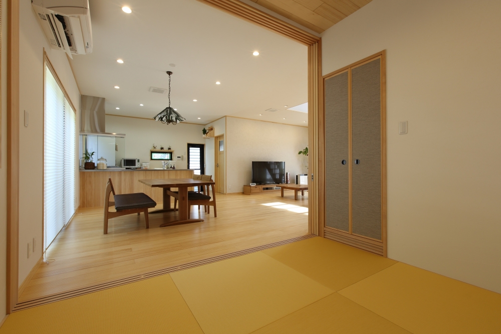 和モダン住宅の畳スペースからLDKを望む空間設計。対面式キッチンとダイニング、リビングが一体となった開放的な間取りで、無垢フローリングと木の質感がやさしく調和。畳との段差を抑えた設計により視線の広がりと動線のスムーズさを実現。奈良の注文住宅における、和室とLDKを美しくつなぐホテルライクで上質な住まいの事例。