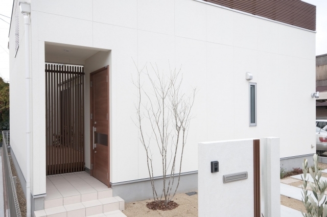 　和モダン住宅の外観と玄関アプローチ。白い塗り壁のシンプルなファサードに木製ルーバーと木製玄関ドアを組み合わせ、水平ラインが美しく整うデザイン。奥行きのある玄関ポーチと格子が外部からの視線をやわらかく遮りながら、光と風を取り込む設計。植栽と砂利敷きの外構が静けさと上質さを演出し、奈良で注文住宅・デザイナーズ住宅・ホテルライクな家を検討する方に向けたプライバシー性と開放感を両立した住まい。