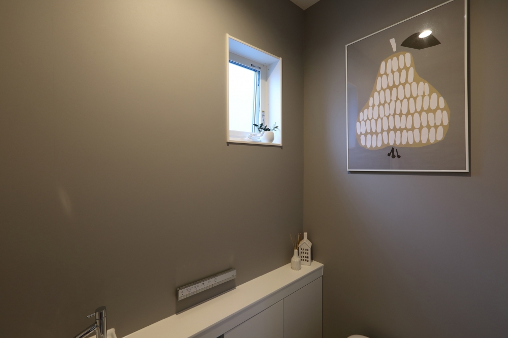 グレートーンの壁面にアートを配したシンプルで上質なトイレ空間。タンクレストイレとフラットなカウンター収納により生活感を抑え、小窓からの自然光でやわらかな明るさと抜け感を演出。ミニマルな設計と素材の質感が調和した、ホテルライクで洗練されたデザイナーズ住宅のトイレインテリア。