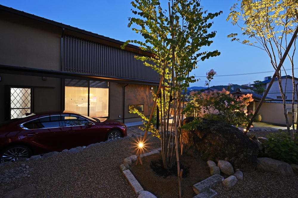 奈良県の和モダン注文住宅の夜景外観。ライトアップされた庭と植栽、石組みの外構と建物が調和し、木格子と大開口の窓から漏れる灯りが陰影を演出。外構と建築が一体となり、光と影で静けさと上質な暮らしを実現した住宅事例。