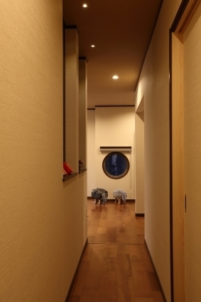 和モダン住宅の廊下から玄関ホールを望む空間設計。直線的に伸びる廊下の先に丸窓を配置し、視線の抜けと奥行きを演出。壁面ニッチや間接照明が陰影を生み、無垢フローリングと落ち着いた内装材が上質な静けさを形成する。動線と視線設計を融合させ、ホテルライクで洗練された暮らしを実現する住宅デザインの一例。