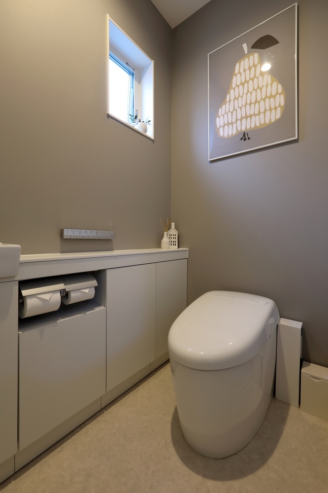 グレートーンのアクセントクロスとアートを組み合わせたホテルライクなトイレ空間。タンクレストイレとフラットなカウンター収納により、清掃性と美しさを両立。壁面収納にトイレットペーパーを収め生活感を抑えつつ、小窓からの自然光で明るさと抜け感を確保。限られた空間を上質に整えた、デザイナーズ住宅の洗練されたトイレ設計。