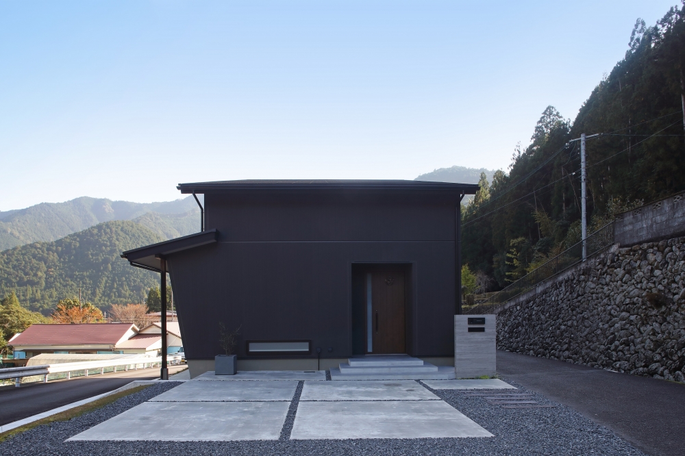 奈良の自然環境に調和する和モダン住宅の外観。黒を基調としたシンプルで重厚感のあるファサードに、深い軒と水平ラインが美しい落ち着いたデザイン。周囲の山並みや石積みの景観と調和しながら、プライバシーを確保した静かな住まいを実現。ミニマルでホテルライクな外観と、自然と共に暮らす設計思想を体現した、注文住宅・デザイナーズ住宅を検討する方に向けた上質な住空間。