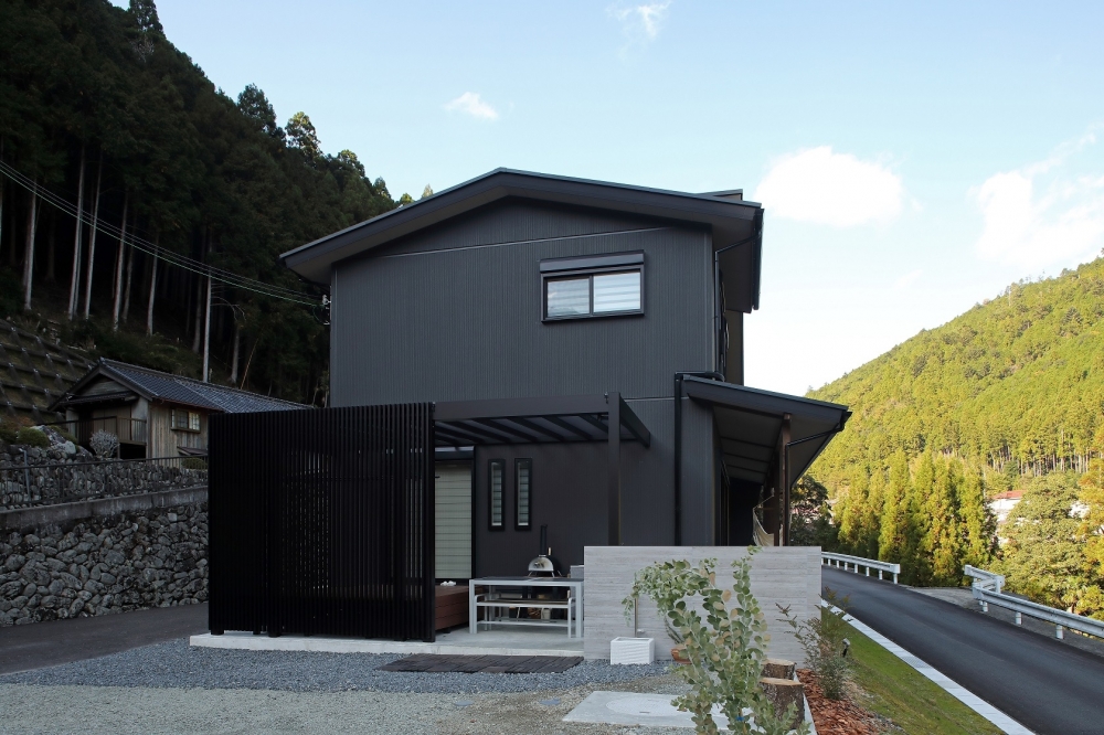 奈良の自然に囲まれた和モダン住宅の外観とアウトドアリビング空間。黒を基調としたシンプルで重厚感のあるデザインに、格子スクリーンが外部からの視線をコントロール。半屋外のテラスにはアウトドアキッチンを備え、グランピングのように自然を楽しめる設計。深い軒と水平ラインが美しく、プライバシーと開放感を両立した注文住宅・デザイナーズ住宅・ホテルライクな住まい。