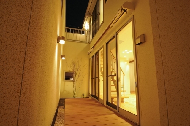 奈良の注文住宅における中庭テラス空間の夜景。外部からの視線を遮る壁に囲まれたプライベートな中庭にウッドデッキを設け、大開口の掃き出し窓を通して室内とシームレスにつながる設計。間接照明と外壁照明が陰影を生み出し、和モダンとホテルライクな上質空間を両立した住まいの実例。