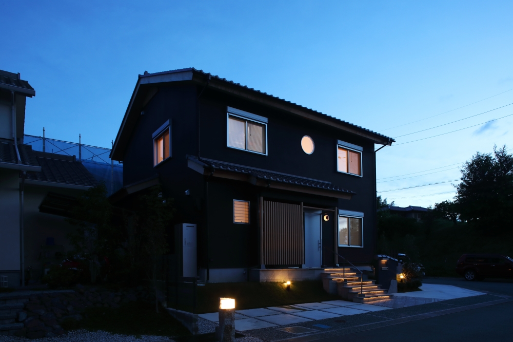 夕景の中に静かに浮かび上がる、黒を基調とした和モダン住宅の夜景外観。深い軒と木格子、丸窓がやわらかな灯りに包まれ、日本建築らしい陰影の美しさを際立たせている。階段アプローチの間接照明と外構照明が、ホテルライクな上質感と安心感を演出し、昼とは異なる落ち着いた表情を生み出す設計。和風住宅・純和風の家・黒い外観の家・和モダン住宅・デザイナーズ住宅・高級感のある注文住宅を求める家づくり検討者へ向けた住まい事例。建築・照明・外構計画を一体で整えた、夜の景観まで美しい和モダン住宅。