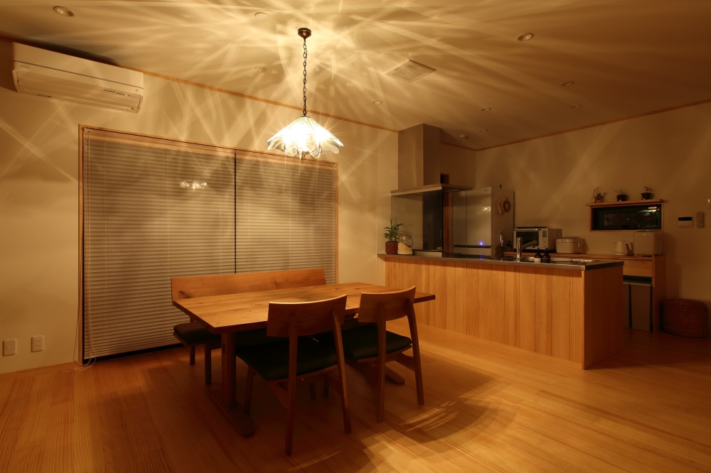 奈良の注文住宅における和モダンのダイニングキッチン空間。無垢材のダイニングテーブルと対面キッチンを一体化し、ペンダント照明の光が天井と壁に広がる照明計画を採用。温かみのある間接光が空間全体を包み込み、ホテルライクで上質な食事時間を実現する住宅デザイン事例。