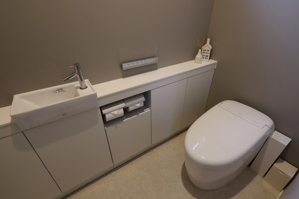 タンクレストイレと一体型手洗いカウンター収納を備えた、ミニマルで洗練されたトイレ空間。グレージュの壁と白の設備が調和し、生活感を抑えながら収納力と機能性を確保。ホテルライクで上質なデザイナーズ住宅の水まわり設計事例。