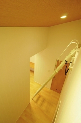 奈良の注文住宅におけるロフトアクセス空間の設計事例。天井収納へとつながる可動式階段を設けた機能的な動線計画で、限られた空間を有効活用。木質フローリングとやわらかな照明が温かみのある室内環境をつくり、和モダンとホテルライクな快適性を兼ね備えた住まい。