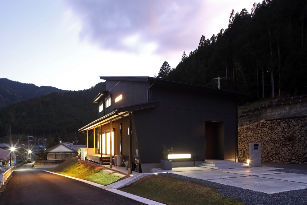 奈良の山間地域に建つ和モダン住宅の夜の外観。黒を基調とした外壁と水平ラインの美しいフォルムが景観に溶け込み、軒下の間接照明と室内の光が温かくにじむ設計。自然と調和しながら上質な暮らしを実現するホテルライクなデザイナーズ住宅事例。