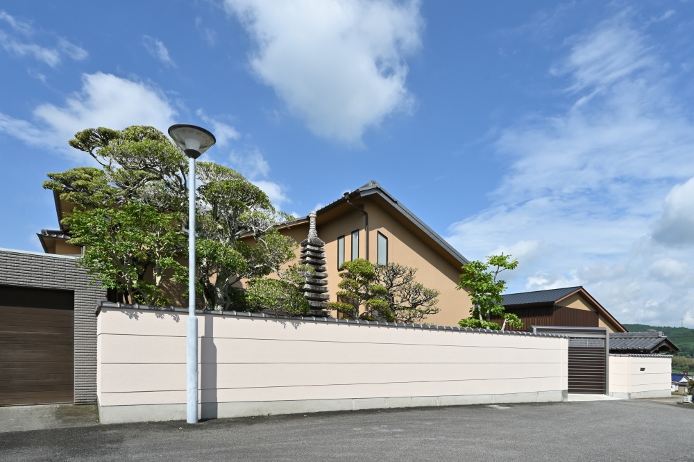 奈良県の和モダン注文住宅の外観と外構。塀で囲われたプライベートな庭に、手入れされた植栽と石塔が配され、落ち着いた佇まいを形成。深い軒と伝統的な屋根形状が特徴で、周辺環境と調和しながら静けさと品格ある暮らしを実現した住宅事例。