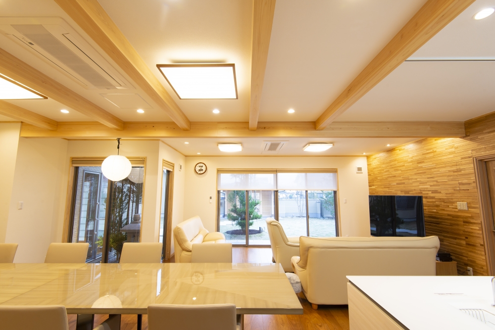 奈良県のやまぐち建築設計室が設計した和モダン住宅のLDK全景。無垢材の梁を現した天井と木質アクセントウォールが特徴のリビングに、ダイニングとアイランドキッチンを一体化した間取り設計。大開口サッシから庭へとつながる開放的な空間構成で、採光・通風・動線を最適化。自然素材の温もりとホテルライクな上質感を兼ね備えた高級注文住宅の実例。