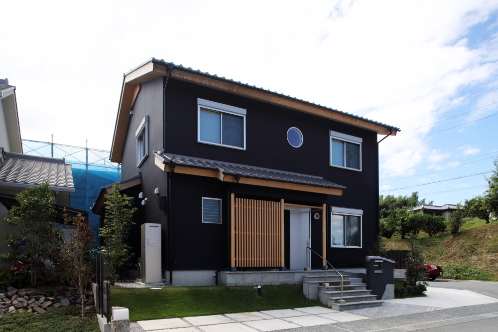 黒を基調とした和モダン住宅の外観と、傾斜地を活かした芝生の外構計画。深い軒と木格子、丸窓が日本建築らしい趣を演出し、落ち着いた佇まいの中に現代的な洗練を感じさせるデザイン。玄関前には来客時にもゆとりを感じるフリー空間を設け、建築と外構を一体で設計することで、暮らしに開放感と品格を与えている。和風住宅・純和風の家・和モダン住宅・黒い外観の家・ホテルライクな家・デザイナーズ住宅を求める家づくり検討者へ向けた、自然と調和する上質な注文住宅事例。