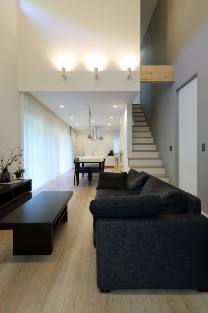 和モダン住宅の吹き抜けリビングとLDK空間。白を基調としたシンプルモダンなインテリアに、木質フローリングとブラックの家具がコントラストを形成。大開口のカーテン越しに自然光がやわらかく広がり、リビングからダイニング・キッチンまで視線が抜ける開放的な間取り。階段と吹き抜けにより上下階がゆるやかにつながる、注文住宅・デザイナーズ住宅・ホテルライクな上質空間。
