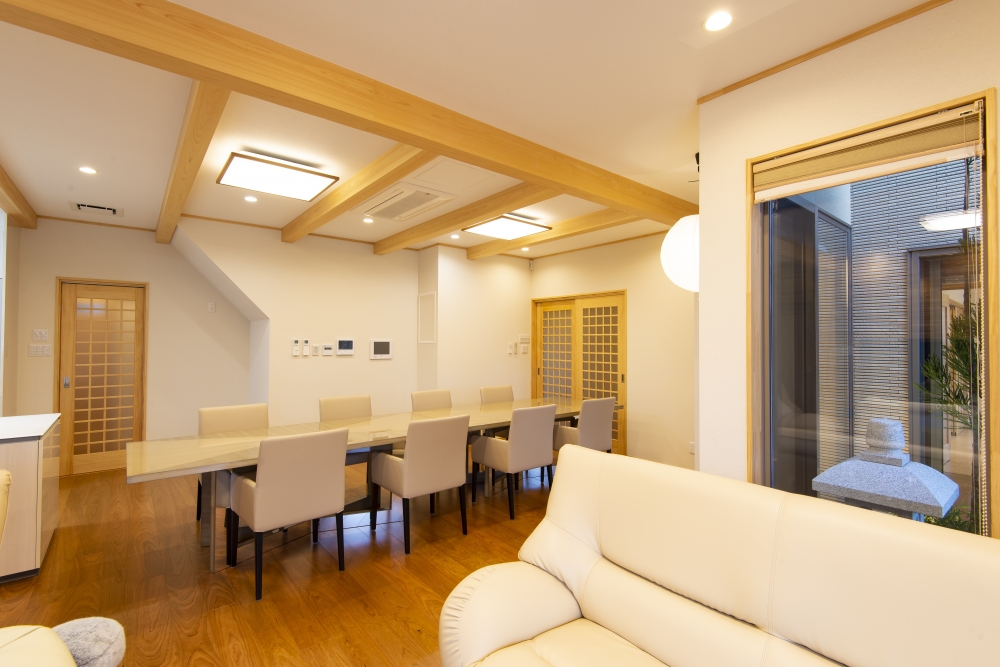 奈良県のやまぐち建築設計室が設計した和モダン住宅のダイニング空間。無垢材の梁を現した天井と上質な木質フローリング、長尺ダイニングテーブルを中心に据えたレイアウト設計。障子格子の引き戸や間接的に庭とつながる開口部により、採光と視線の抜けを確保。リビングと連続する開放的なLDKで、家族の時間と来客対応の両立を実現した高級注文住宅の実例。