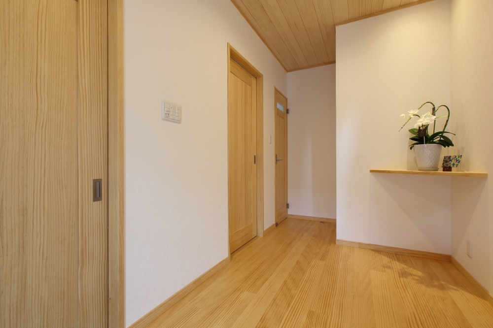 和モダン住宅の玄関ホールから続く廊下空間。無垢材の床と木天井、木製建具が統一された温もりある内装で、シンプルながら上質な設え。壁面の飾り棚が空間に余白と奥行きを与え、生活動線を整えながら落ち着きのあるホテルライクな住まいを実現。自然素材と整った動線計画が調和した注文住宅の好例。