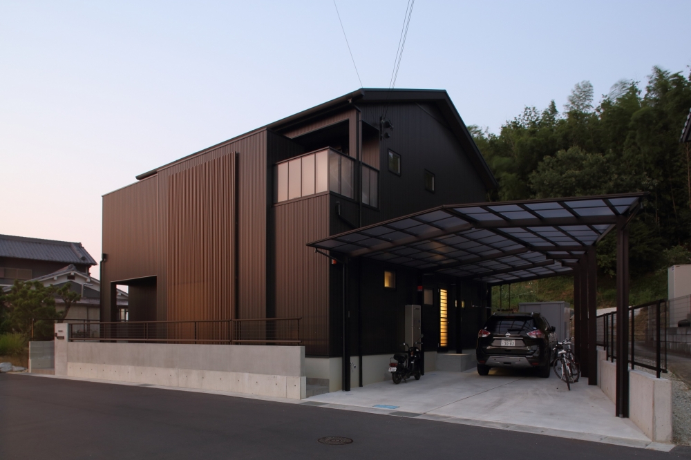 奈良の注文住宅における和モダン住宅の外観デザイン。黒の外壁と縦格子を組み合わせたシンプルモダンなファサードに、機能的なカーポートと駐車スペースを一体計画。プライバシーを確保しながら、暮らしやすさと動線を重視した設計。夕景に映える外構と建物のバランスが、ホテルライクで上質な住まいを演出する高級住宅事例。