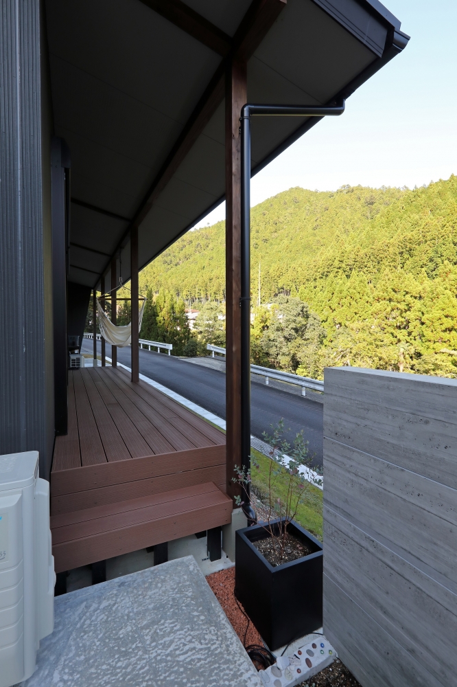 奈良の自然を望む和モダン住宅のウッドデッキと半屋外空間。黒を基調とした外壁と木製柱、深い軒に守られたテラスが特徴で、ハンモックを設えたアウトドアリビングとして活用できる設計。コンクリート塀が外部からの視線を遮りつつ、山並みの景色や風、光を取り込むバランスの取れた空間構成。注文住宅・デザイナーズ住宅・ホテルライクな住まいを求める方に向けた上質な住環境。