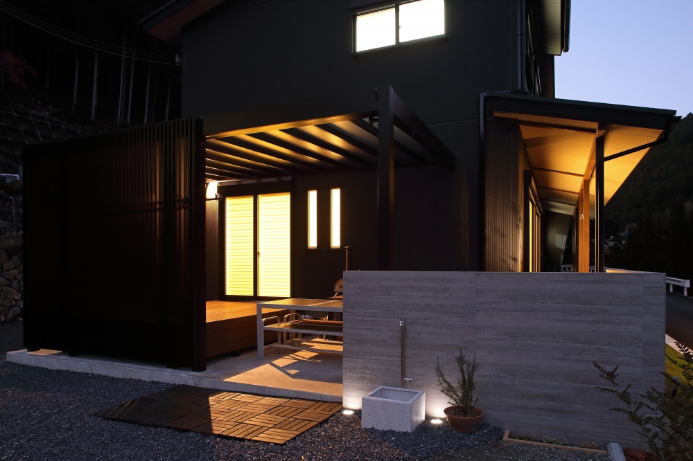 夜の照明に包まれた和モダン住宅の外観。黒を基調とした外壁と木格子が外部からの視線をやわらかく遮り、内側には暖かな光が広がる設計。中庭やデッキ空間と連続する半屋外テラスが、ホテルライクで上質な暮らしを演出するデザイナーズ住宅事例。
