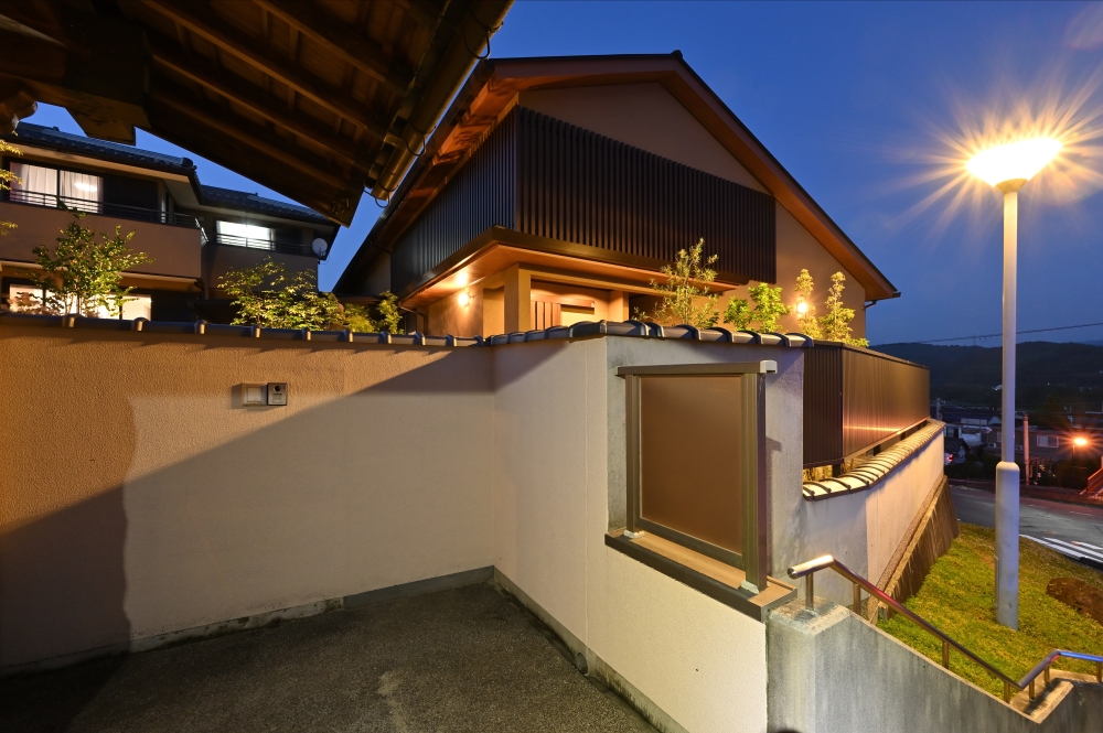奈良県の和モダン注文住宅の夜景外観。塀で囲まれた外構と木格子のファサードに、温かみのある照明が陰影を生み出す住まい。深い軒と外壁の質感が調和し、外からの視線を遮りながら静けさと品格ある暮らしを実現した住宅事例。