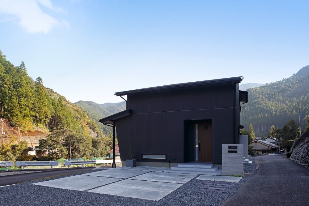 奈良の山並みに囲まれた自然環境に調和する和モダン住宅の外観。黒を基調としたシンプルで重厚感のあるファサードに、深い軒とシャープな水平ラインが美しいデザイン。周囲の景観と呼応しながらプライバシーを確保し、静けさと開放感を両立。コンクリートと砂利敷きの外構がミニマルな美しさを引き立て、注文住宅・デザイナーズ住宅・ホテルライクな住まいを求める方に適した上質な住空間。