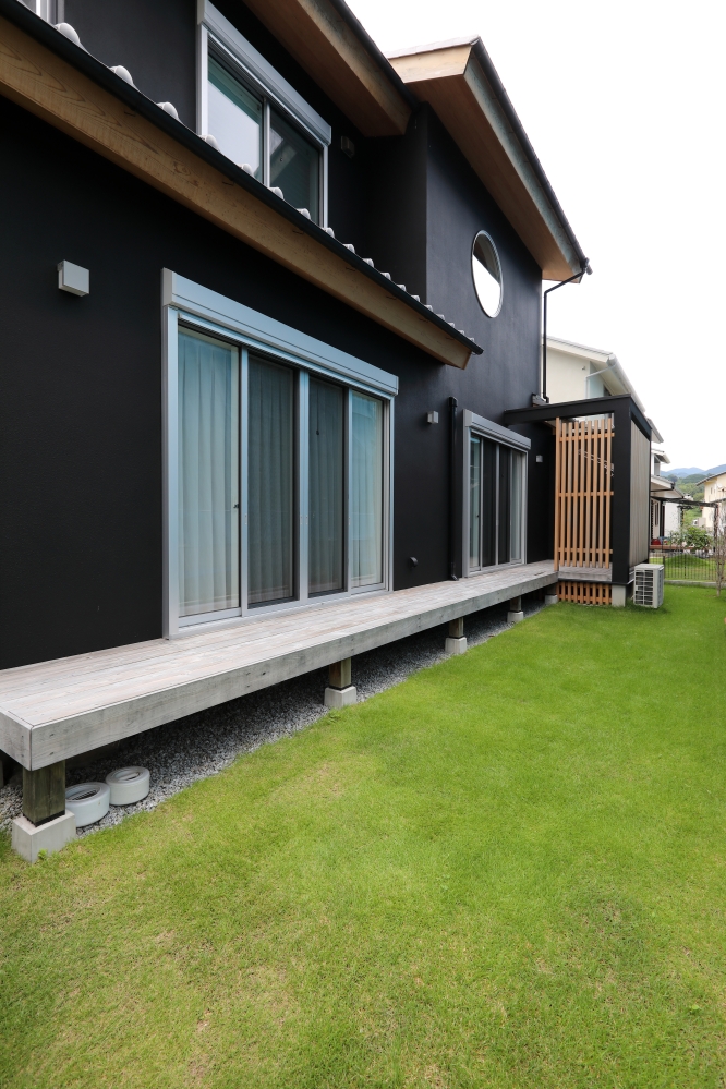 黒を基調とした和モダン住宅の外観と、LDKからフラットに繋がる天然木のウッドデッキ、芝生の庭が一体となった住まい。深い軒と丸窓、日本建築らしい水平ラインが落ち着いた景観を生み出し、外と内を緩やかに繋ぐ設計が特徴。リビングから庭へ視線と動線が自然に広がり、家族時間やアウトドアリビングを楽しめる空間構成。和風住宅・純和風の家・和モダン住宅・ホテルライクな家・黒い外観の家・デザイナーズ住宅を求める家づくり検討者へ向けた、自然と共に暮らす上質な注文住宅事例。