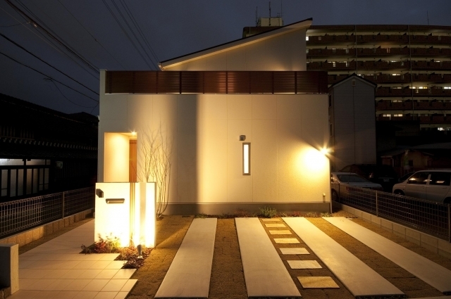 奈良の注文住宅における和モダン外観と外構デザインの夜景。白い外壁と片流れ屋根のシンプルなフォルムに、格子と間接照明を組み合わせ、プライバシーを確保しながら上質な光の演出を実現。アプローチと駐車スペースを一体的に設計し、ホテルライクで落ち着きのある住まいを表現した住宅事例。
