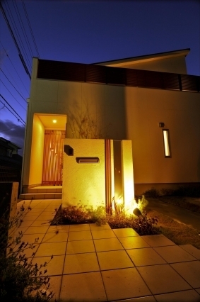 奈良の注文住宅における和モダンの外構・玄関アプローチの夜景。白い外壁と片流れ屋根のシンプルな外観に、間接照明と植栽照明を組み合わせ、格子の玄関がやわらかく浮かび上がる設計。光と影のコントラストによりプライバシーと奥行きを演出し、帰宅時に心地よさを感じるホテルライクな住宅デザイン事例。