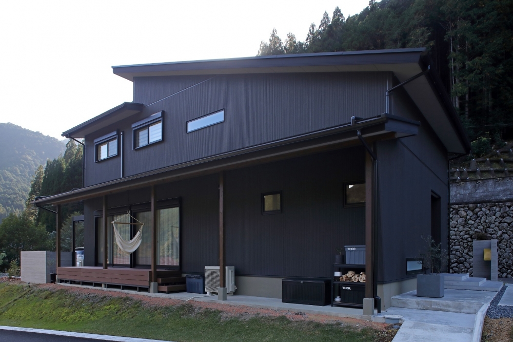 　奈良の自然に囲まれた和モダン住宅の外観とウッドデッキ空間。黒を基調とした落ち着きのある外観に、深い軒と水平ラインが美しく伸びるデザイン。屋根に守られた半屋外のウッドデッキは、ハンモックやアウトドアリビングとして活用でき、内と外を緩やかにつなぐ設計。プライバシーを確保しながら自然を取り込む、注文住宅・デザイナーズ住宅・ホテルライクな住まいを求める方に向けた上質な住空間。