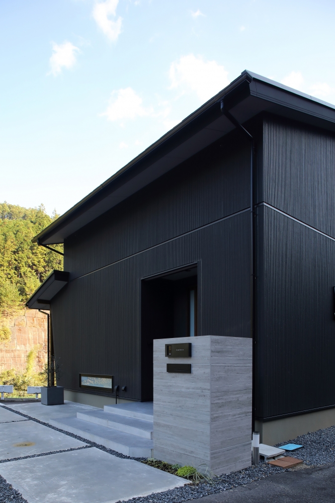 奈良の自然環境に調和する和モダン住宅の外観デザイン。黒を基調とした外壁に繊細な縦ラインのテクスチャが施され、深い軒とシャープな水平ラインが美しいファサード。コンクリート門柱とシンプルなアプローチが外観の重厚感と品格を引き立て、視線をコントロールしながらプライバシーを確保。注文住宅・デザイナーズ住宅・ホテルライクな住まいを求める方に向けた、静けさと上質さを兼ね備えた住空間。
