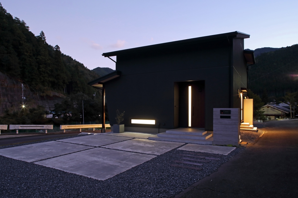 奈良の自然環境に佇む黒い外観の和モダン住宅の夜景。シンプルで直線的なフォルムと最小限の開口計画によりプライバシーを確保しつつ、玄関や外構の間接照明が上質な光の陰影を演出。ホテルライクで洗練されたデザイナーズ住宅の外観事例。