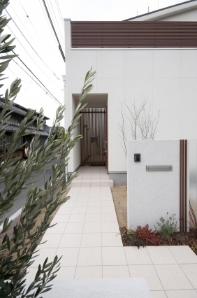 和モダン住宅の玄関アプローチと外観デザイン。白い塗り壁のシンプルなファサードに木製ルーバーと水平ラインを組み合わせ、落ち着きのある外観を形成。タイル敷きのアプローチが奥行きを生み、格子戸のある玄関へと視線を導く設計。外構の植栽と門柱がプライバシー性と美観を高め、奈良で注文住宅・デザイナーズ住宅・ホテルライクな住まいを検討する方に向けた、静けさと上質さを兼ね備えた住空間。