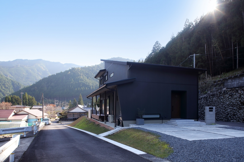奈良の山並みに囲まれた自然環境に建つ和モダン住宅の外観。黒を基調としたシンプルで重厚感のあるデザインに、深い軒と水平ラインが調和し、周囲の景観と美しく馴染む佇まい。道路からのアプローチと外構計画が敷地との高低差を活かしながら、プライバシーと開放感を両立。注文住宅・デザイナーズ住宅・ホテルライクな住まいを求める方に向けた、自然と共に暮らす上質な住空間。