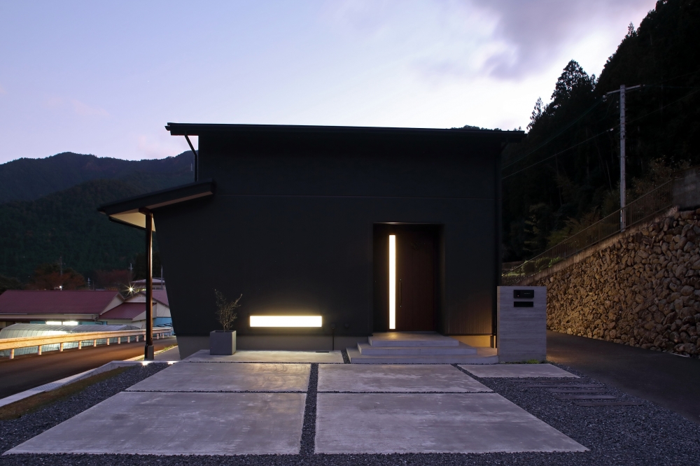 黒い外壁で構成された和モダン住宅の夜の正面外観。最小限の開口と直線的なフォルムによりプライバシーを確保しつつ、玄関の縦スリット窓と外構照明が柔らかな光を演出。ホテルライクで洗練されたデザイナーズ住宅の外観設計事例。