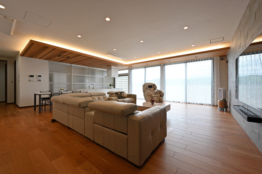 和モダン住宅の開放的なリビングダイニング空間。無垢フローリングと木目天井、間接照明による陰影が奥行きと上質感を演出。大開口のカーテン越しに自然光を取り込み、アイランドキッチンからリビングまで視線と動線が一体化した間取り。石調のアクセントウォールと低重心の家具配置により、落ち着きと広がりを両立したホテルライクなLDK設計。自然素材とシンプルな直線美を融合した和モダン住宅の実例。