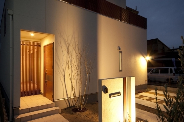 奈良の注文住宅における和モダン外構と玄関アプローチの夜景デザイン。白い外壁と木製玄関ドア、格子スクリーンを組み合わせ、間接照明で陰影を演出。プライバシーを確保しながら上質な迎えの空間を形成し、ホテルライクな静けさと奥行きを感じさせる住宅外観事例。