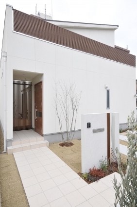 和モダン住宅の外観デザイン。白い塗り壁と水平ラインを強調した木ルーバーが美しいファサード。プライバシーを確保しながら光と風を取り込む設計で、格子扉のある奥行きある玄関アプローチと植栽が上質な暮らしの入口を演出。奈良で注文住宅・デザイナーズ住宅を検討する方に向けた、ホテルライクで静けさを感じる住まい。
