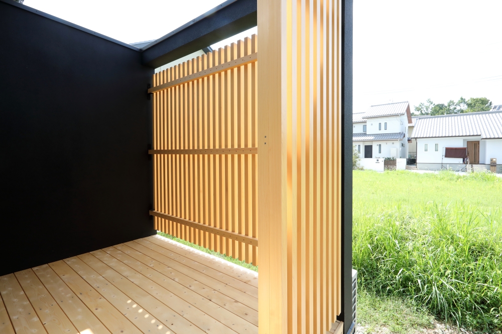 LDKから繋がる天然木のウッドデッキと、木格子で囲われた和モダン住宅の半屋外空間。黒を基調とした外壁に、無垢材の格子と床材が美しく映え、日本建築らしい陰影と落ち着きを演出している。外部からの視線をやわらかく遮りながら、光と風を取り込み、自然と調和した居場所を形成。アウトドアリビングや中間領域として機能する設計で、和風住宅・純和風の家・ホテルライクな家・黒い外観の家・デザイナーズ住宅を求める家づくり検討者へ向けた住まい事例。素材感・余白・内外一体設計を重視した上質な和モダン空間。