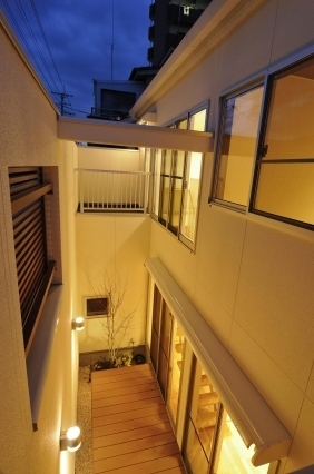 奈良の注文住宅における中庭空間の夜景を上部から見下ろした構図。外壁に囲まれたプライベートな中庭にウッドデッキと植栽を配置し、ブラケットライトと室内照明が陰影と奥行きを演出。上部に開いた空から自然光と風を取り込み、外部と内部を緩やかにつなぐ和モダン・ホテルライクな住まいの設計事例。